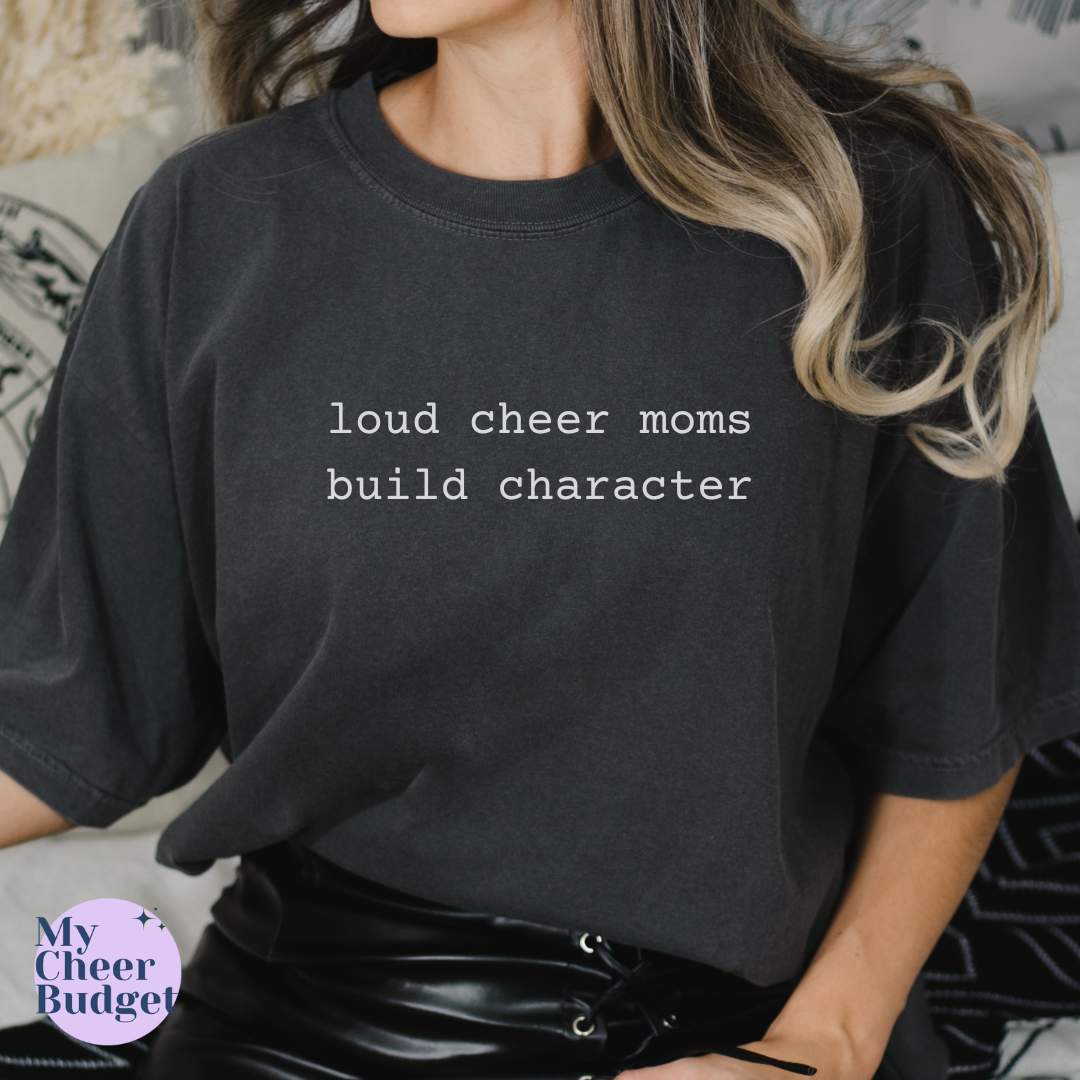Person wearing a black t-shirt with 'loud cheer moms build character' text, sitting on a couch.